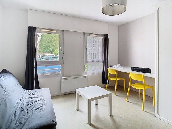 A room with a sofa, a small table, a desk with two yellow chairs, and a window with curtains.