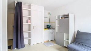 A compact room with a grey sofa, a white wardrobe, a kitchen area with a fridge, and a door leading to another room. - Le Wellington, Arras (Arras)