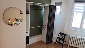 A room with a wooden floor, a white radiator, a black radiator, a black chair, a white wardrobe, a mirror, and a window with curtains. - The Scottman House (Arras)
