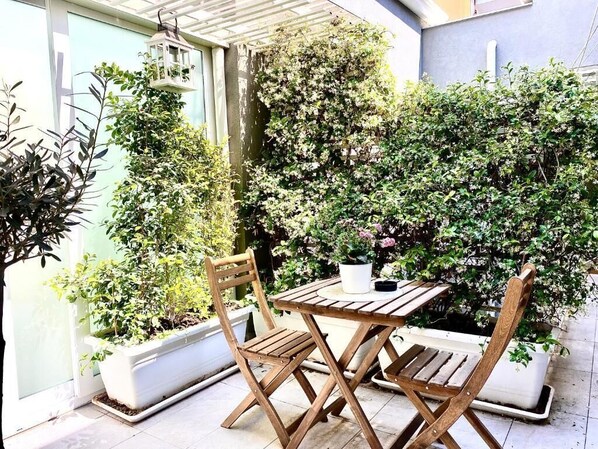 An outdoor seating area with a table and two chairs, surrounded by potted plants and a wall of greenery.