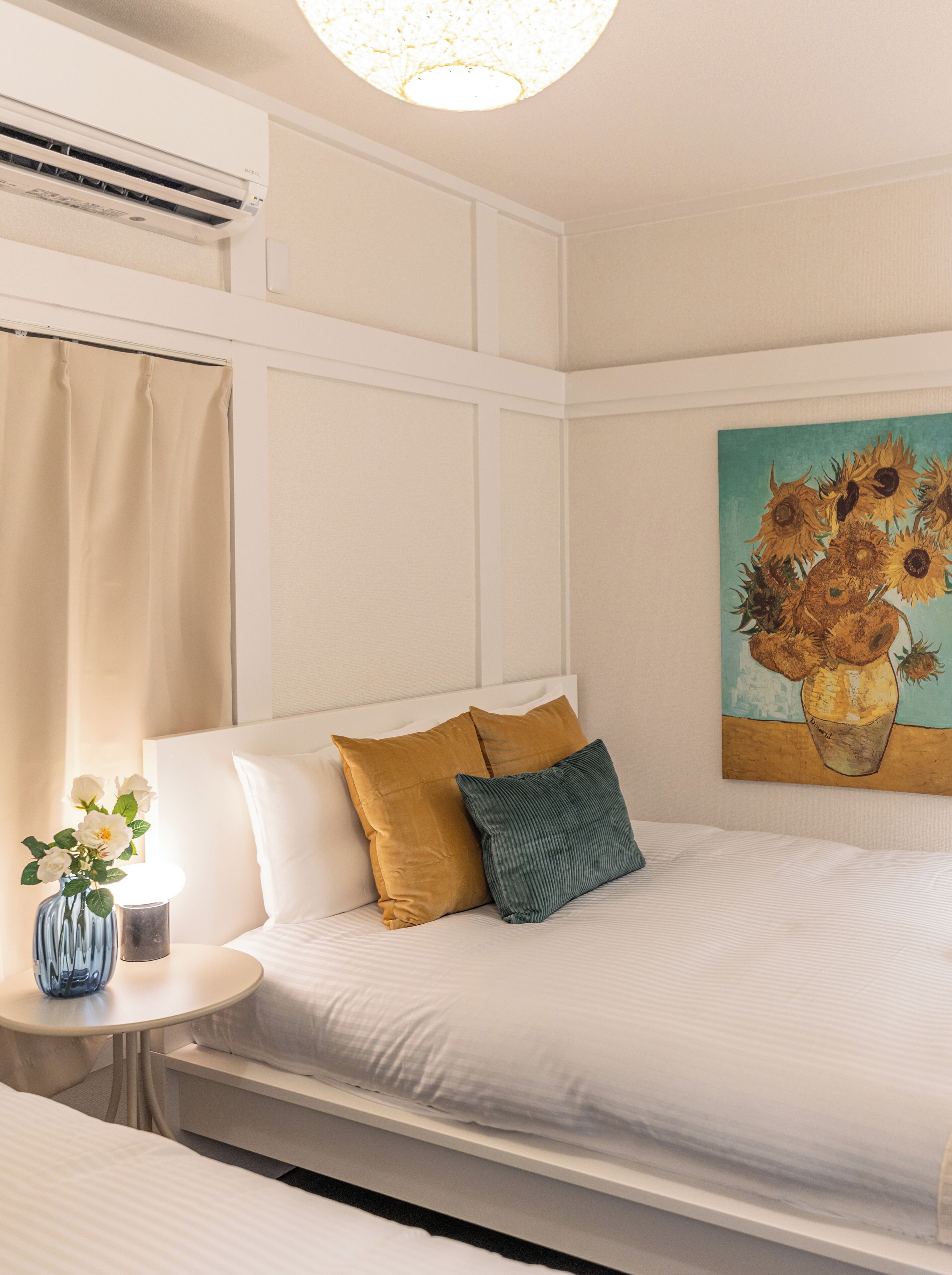 A hotel room with a bed, a bedside table with a vase of flowers, a painting of sunflowers on the wall, and an air conditioning unit above the bed.
