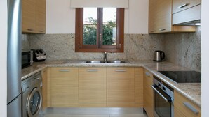 A kitchen with wooden cabinets, a built-in oven, a washing machine, and a window with a view of greenery. - 1 Bedroom Apartment - BH12, Aphrodite Hills Rentals – Apartments (kouklia)