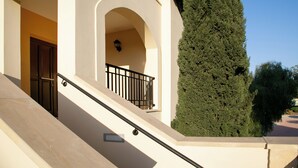 A building with a staircase leading to a balcony, a door, and a tall tree. - 1 Bedroom Apartment - BH12, Aphrodite Hills Rentals – Apartments (kouklia)