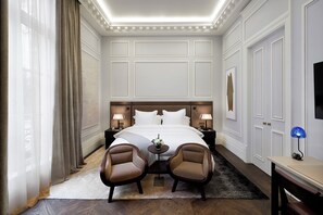 Una habitación de hotel con una cama grande, dos sillones, una mesita pequeña, una mesita de noche y un televisor.