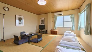 Una habitación con esteras de tatami, un escritorio con silla, una cama con almohadas, un televisor y obras de arte en las paredes.