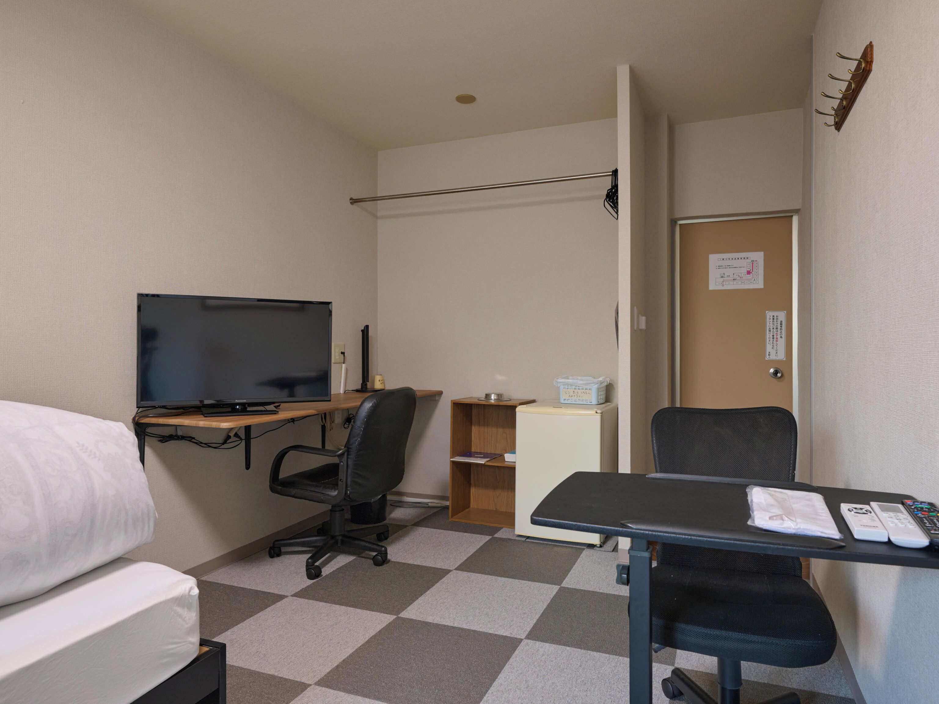 A hotel room with a bed, a desk with a computer, a chair, a television, a refrigerator, and a door with a sign.