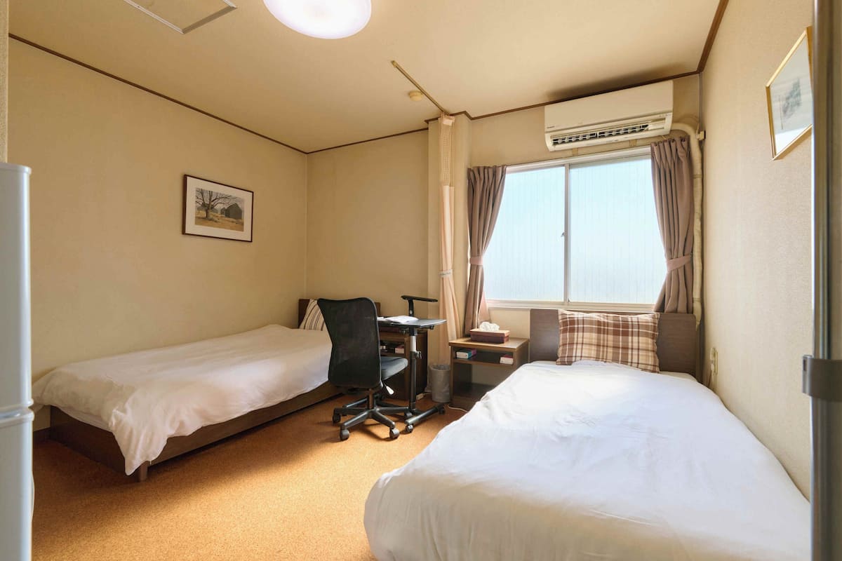 Een hotelkamer met twee eenpersoonsbedden, een bureau met stoel, een raam met gordijnen, een airconditioning en een ingelijst schilderij aan de muur.