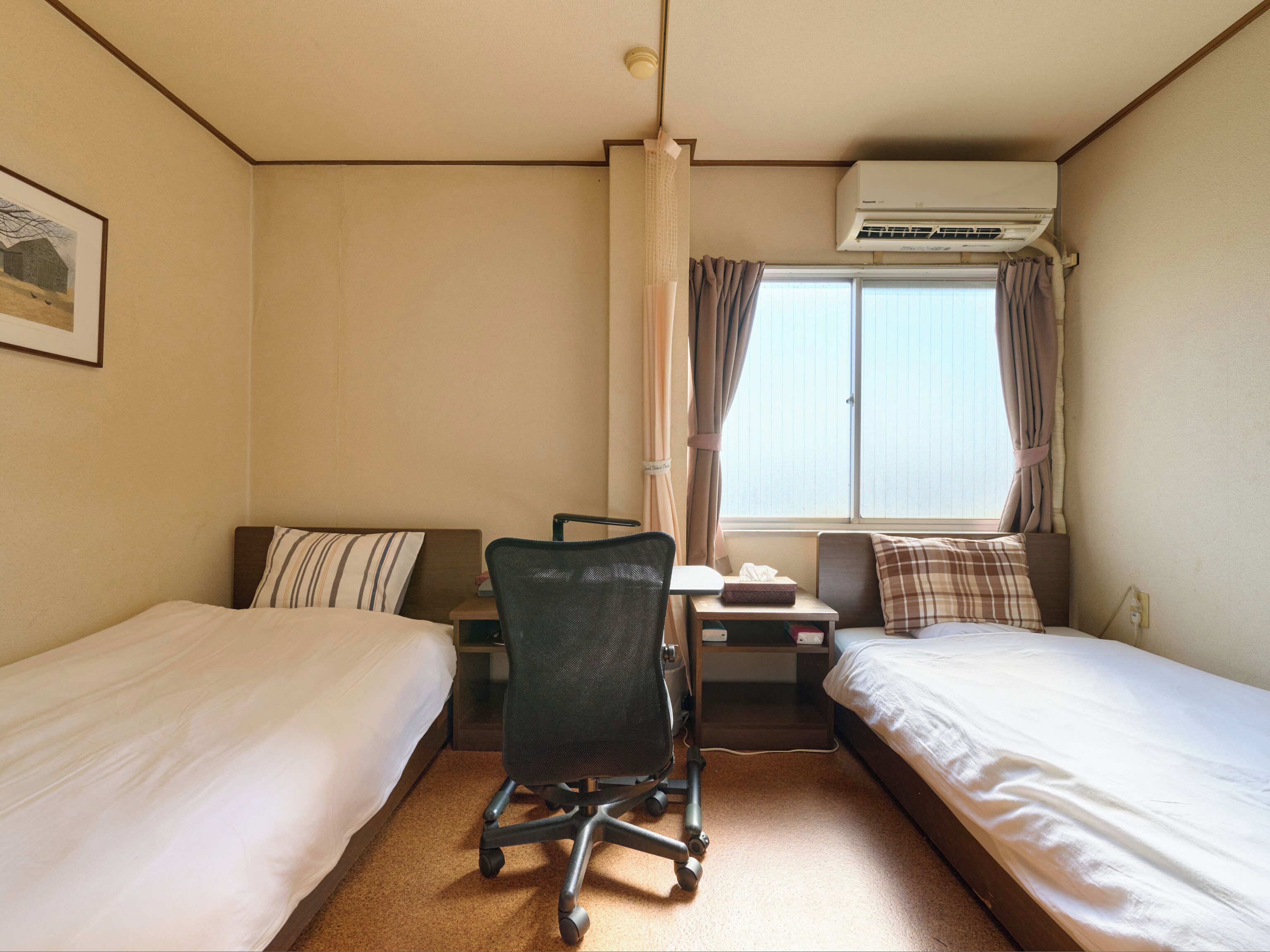 A hotel room with two single beds, a desk with a chair, a window with curtains, and an air conditioning unit.
