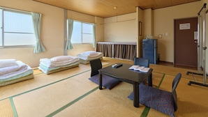 Una habitación con esteras de tatami, una mesa baja, sillas, una cama con almohadas, una ventana con cortinas, una lámpara en el techo y una puerta con un cartel.