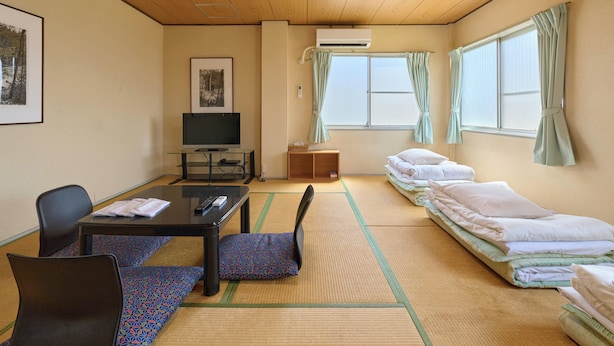 Una habitación con techo de madera, un televisor sobre una mesa de vidrio, esteras de tatami, una cama con almohadas y una ventana con cortinas.
