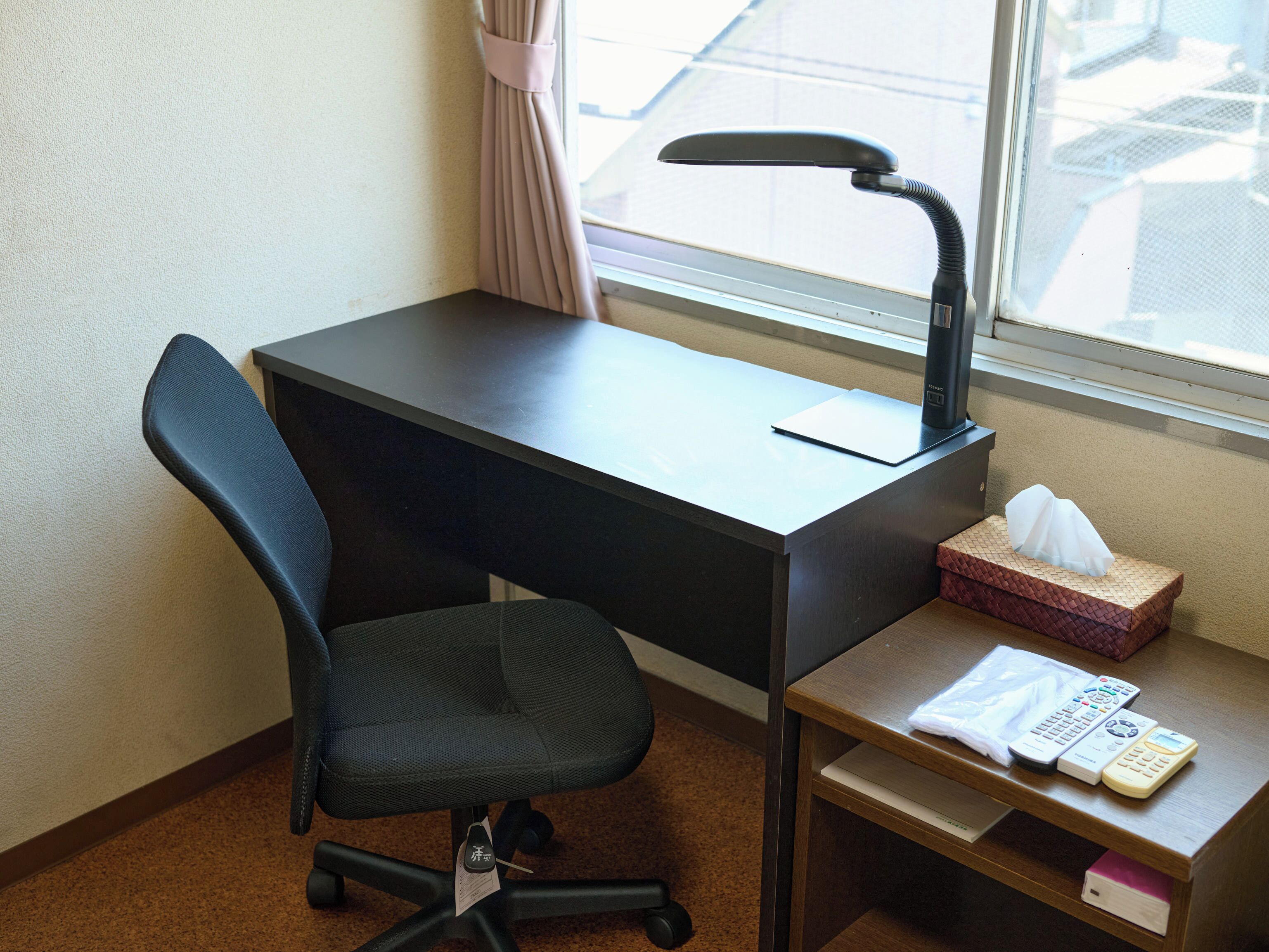 A hotel room with a black desk, a black office chair, a window with a curtain, and a small wooden side table with a tissue box and a remote control.