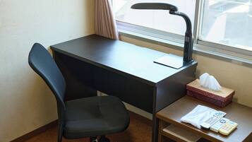 A hotel room with a black desk, a black office chair, a window with a curtain, and a small wooden side table with a tissue box and a remote control.