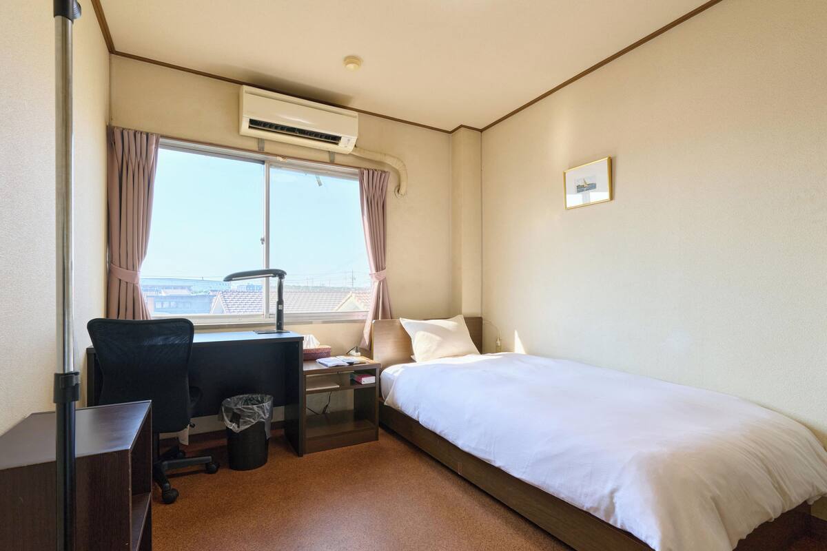Hotelkamer met een enkel bed, een bureau met stoel, een klein tafeltje, een airconditioning en een raam met gordijnen.