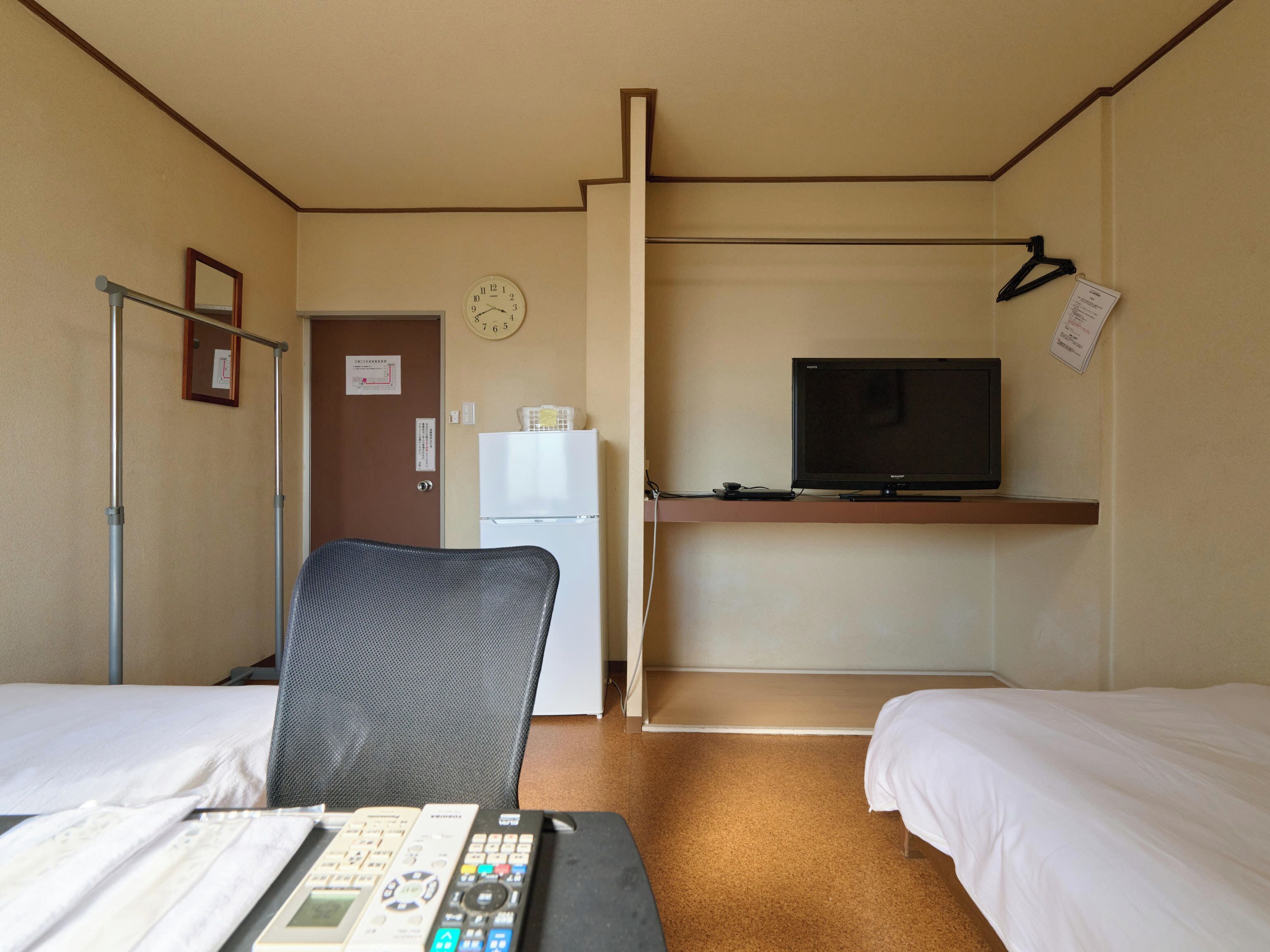 A hotel room with two beds, a television on a stand, a refrigerator, a chair, and a desk with a remote control.