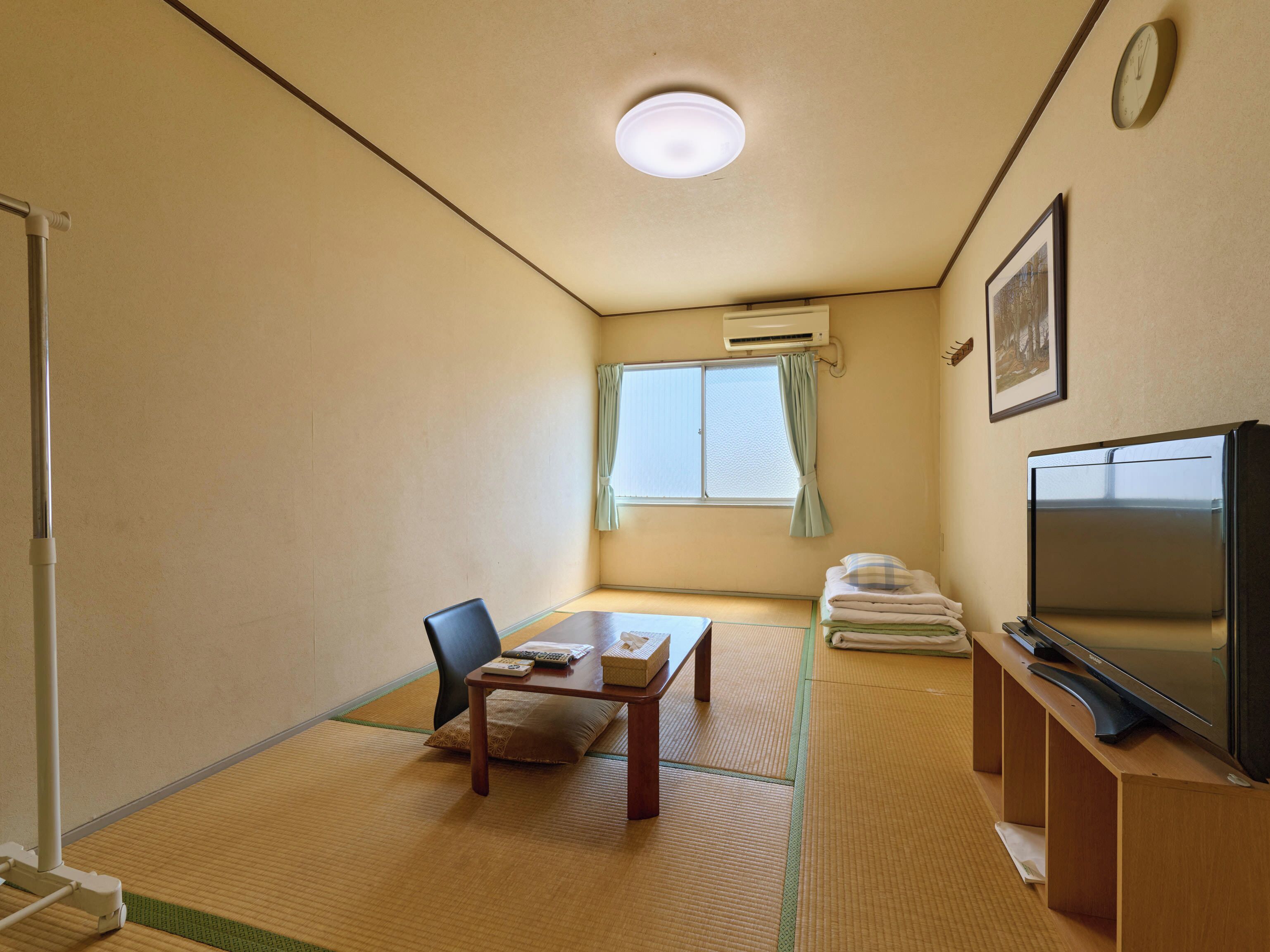 Een kamer met tatami vloerbedekking, een televisie, een klein tafeltje met een kussen, een stoel en een raam met gordijnen.