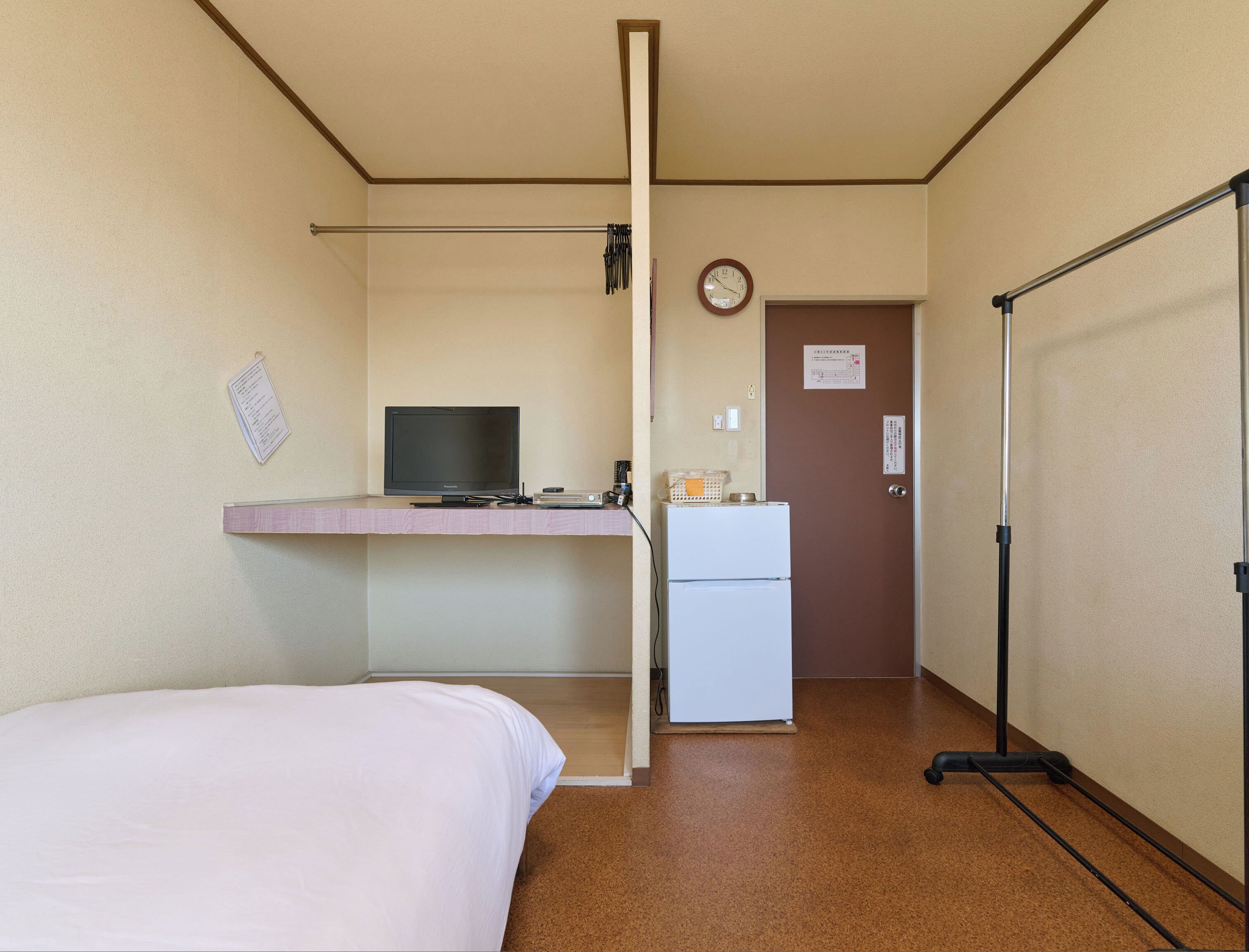 A hotel room with a bed, a desk with a television, a refrigerator, a door with a sign, and a coat rack.