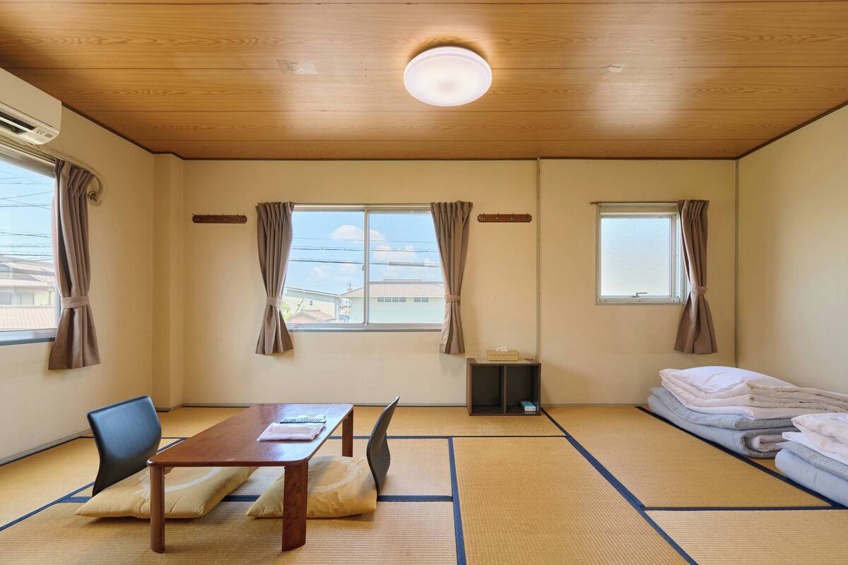 Een kamer met tatami matten, een houten tafel, een stoel, een bed met netjes opgemaakt bedlinnen en een raam met gordijnen.