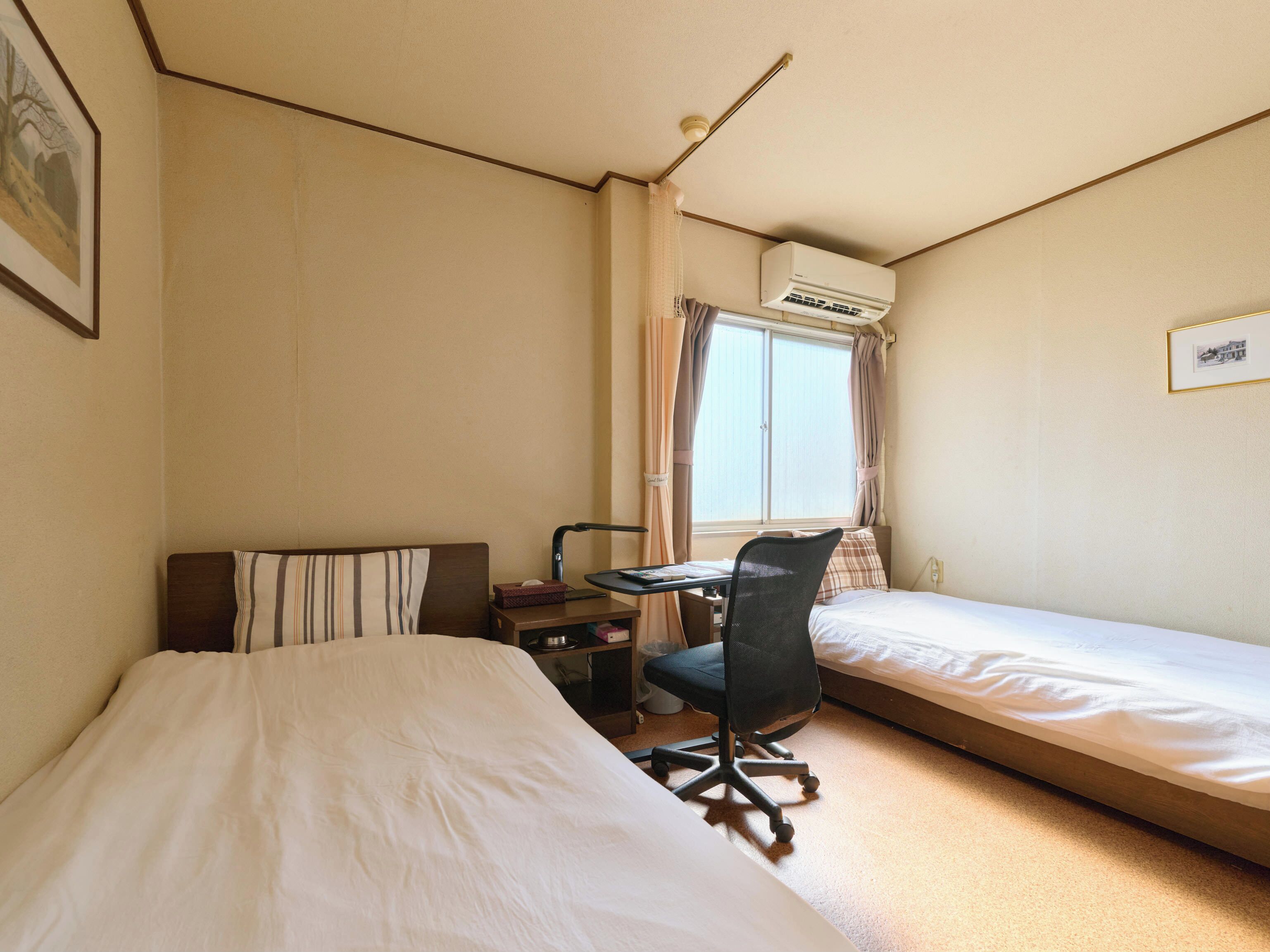 A hotel room with two single beds, a desk with a chair, an air conditioning unit, and a window with curtains.