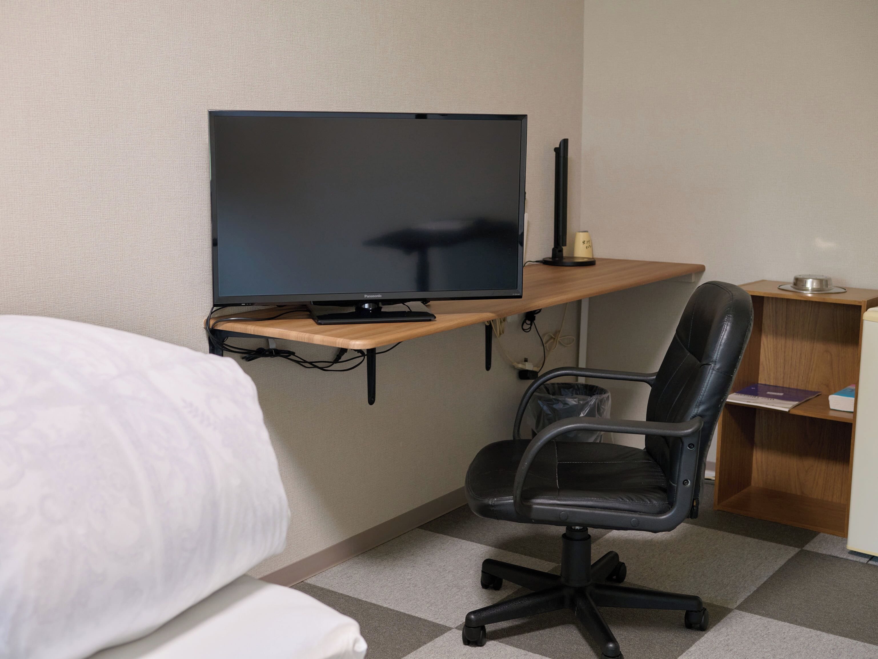 A hotel room with a bed, a desk with a computer monitor, a chair, and a small shelf with books and a cup.