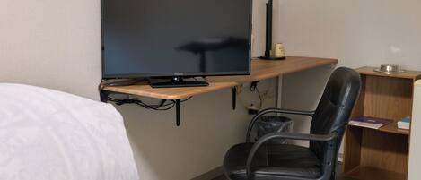 A hotel room with a bed, a desk with a computer monitor, a chair, and a small shelf with books and a cup.