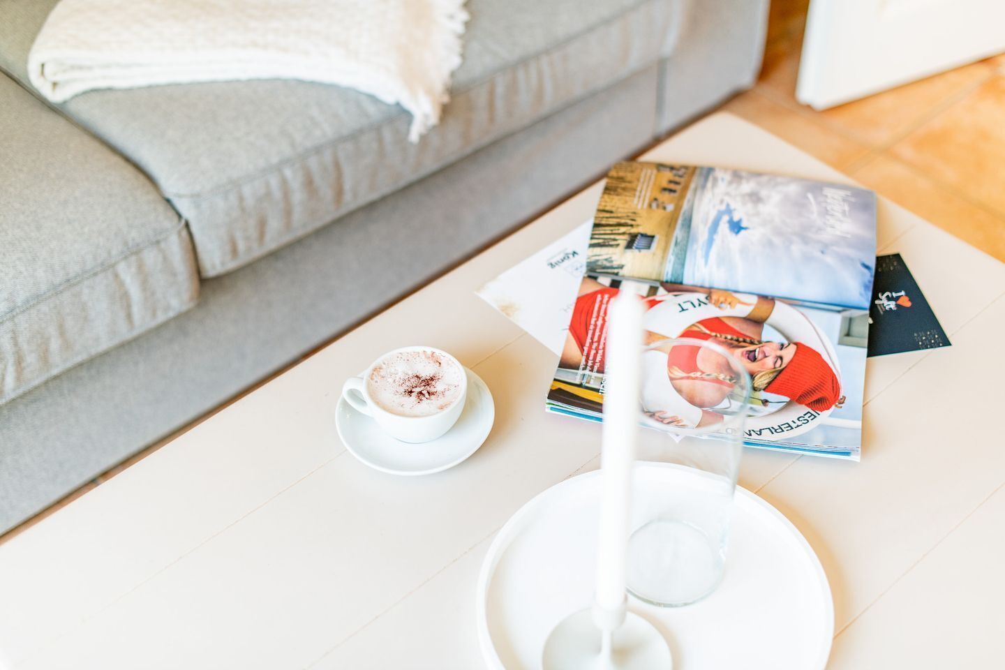 Una taza de café blanca sobre un plato, un vaso de leche y una revista sobre una mesa blanca.