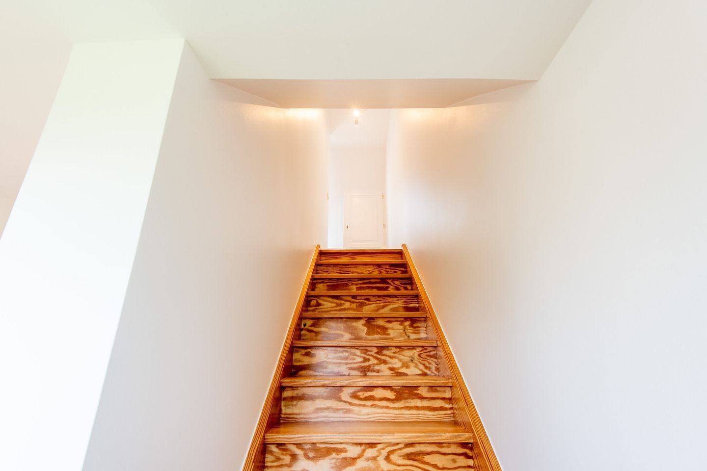 A narrow, wooden staircase with a white wall on one side and a ceiling light at the top.