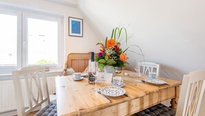 A dining area with a wooden table set for two, white chairs, a vase of flowers, and a bottle of wine. - Huus Lerchenkoje 2 - Huus Lerchenkoje (Sylt)