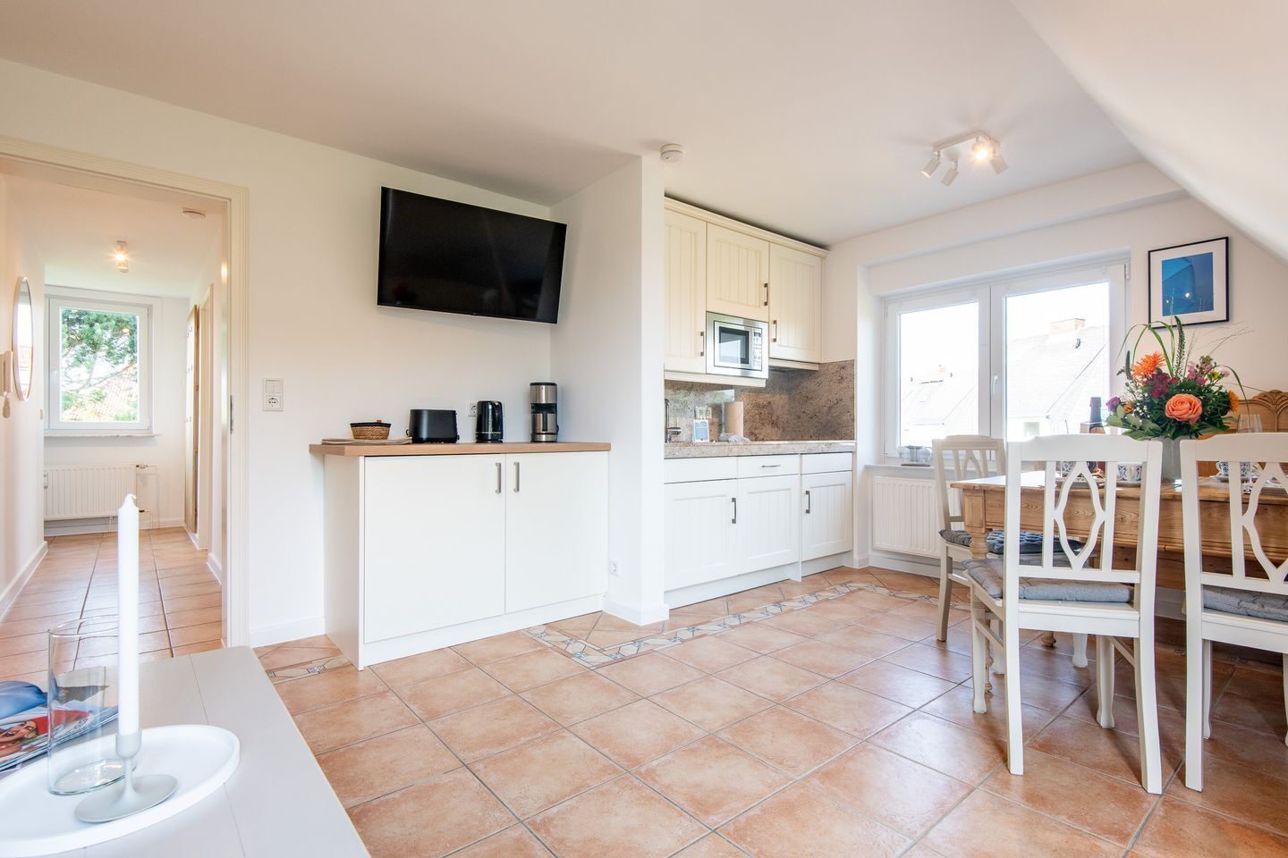 Una cocina moderna con armarios blancos, un televisor de pantalla plana, un microondas y un comedor con mesa y sillas de madera.