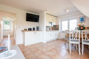 A modern kitchen with white cabinets, a flat-screen TV, a microwave, and a dining area with a wooden table and chairs.