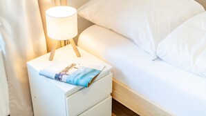 A neatly made bed with white linens and a pillow with a small embroidered logo. A white bedside table with a lamp and a magazine is visible. - Huus Lerchenkoje 2 - Huus Lerchenkoje (Sylt)