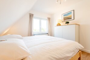 Une chambre à coucher avec un lit, une fenêtre avec des rideaux, une lampe de chevet, un tableau encadré au mur et une petite plante sur une étagère.