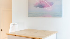 A room with a wooden table, a white wall, and a framed artwork depicting a cloud and ocean. - Huus Lerchenkoje 2 - Huus Lerchenkoje (Sylt)