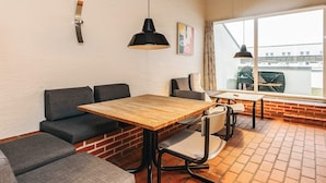 Un intérieur moderne avec une table en bois et des sièges rembourrés de couleur grise, un luminaire suspendu et une grande fenêtre donnant sur des immeubles.