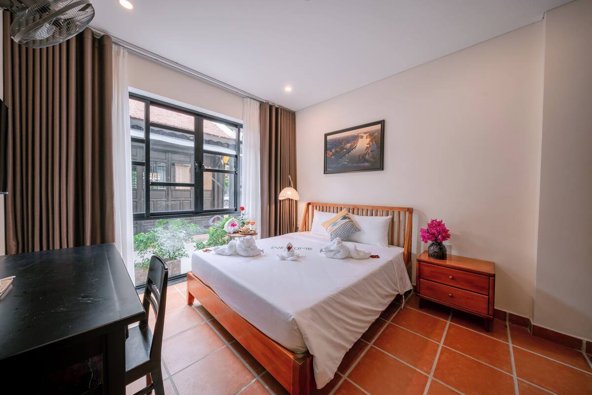 Una habitación de hotel con cama, mesita de noche, lámpara y ventana con cortinas.