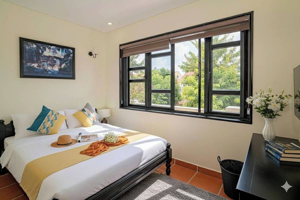 Un dormitorio con cama, ventana con vista a follaje, un cuadro en la pared y una mesita de noche con libros y un jarrón de flores.