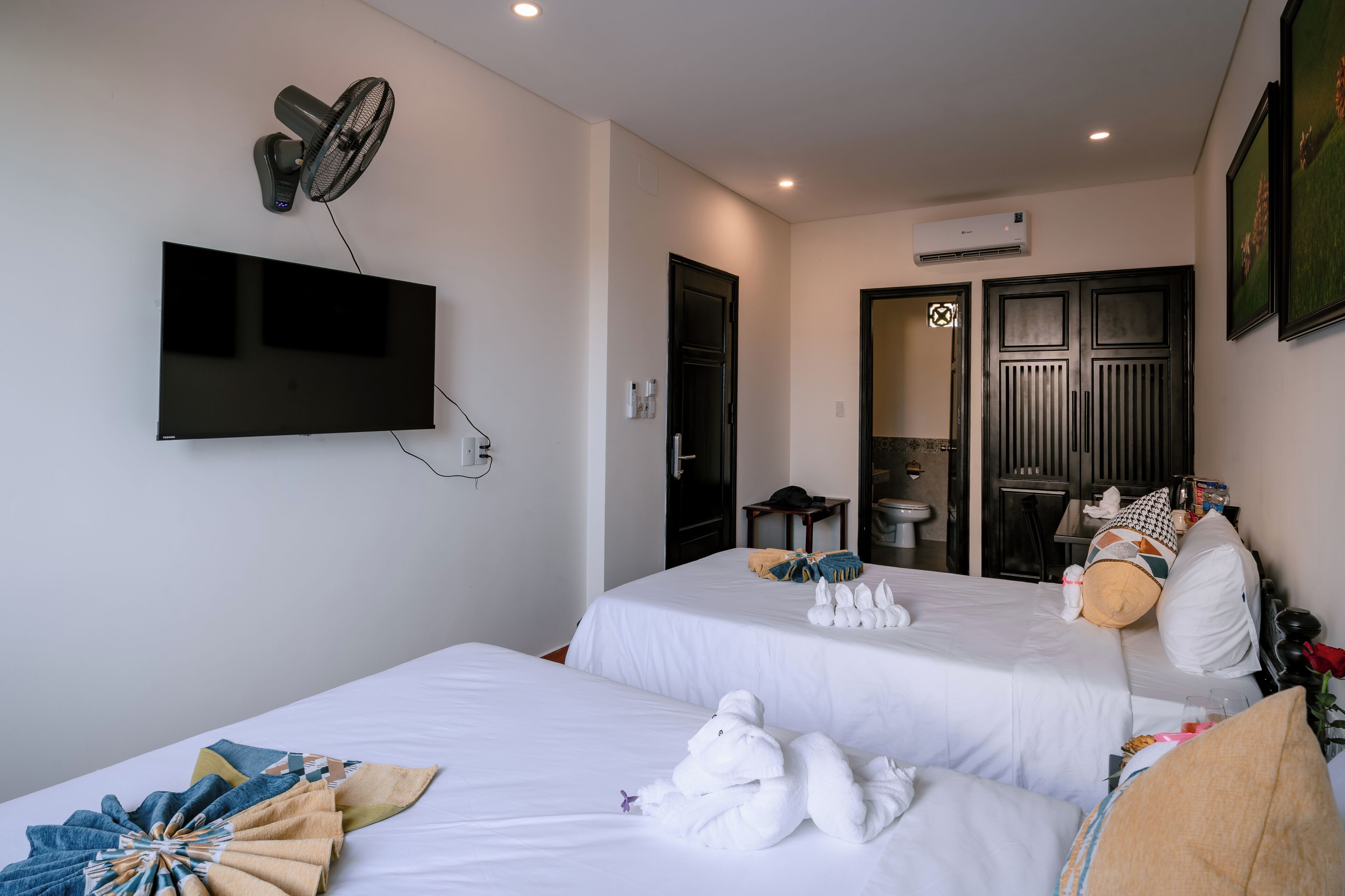A hotel room with two beds, a flat-screen TV, a wall-mounted fan, and an air conditioning unit.