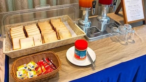 Un buffet de petit-déjeuner comprenant du pain, du beurre, des confitures et un distributeur de jus de fruits.