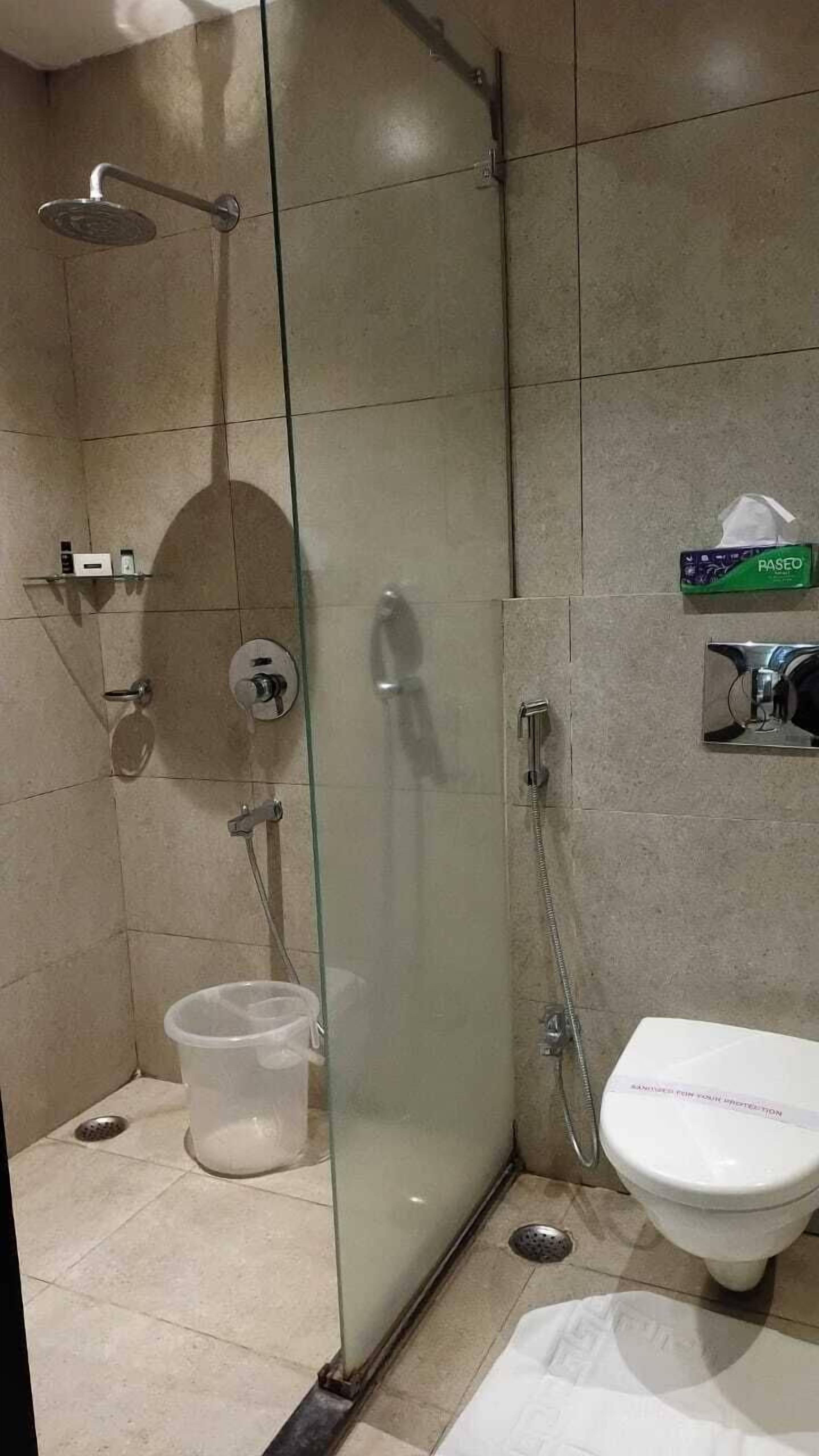 A glass-enclosed shower stall with a handheld showerhead, a handheld showerhead, a toilet, and a small trash bin.
