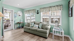 A living room with a green sofa, two windows with white shutters, a small table, and a door leading outside.