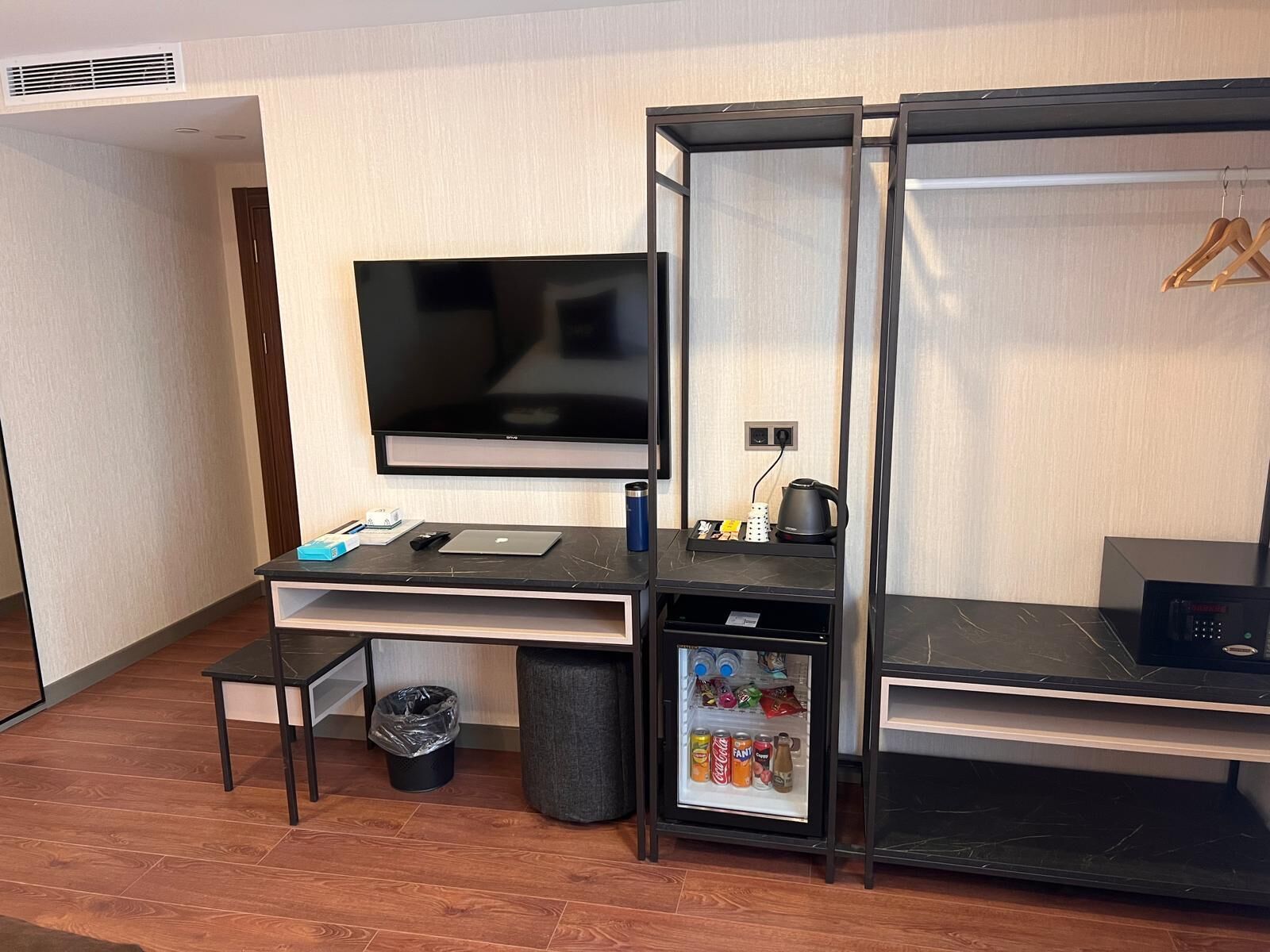 Ein kleines Hotelzimmer mit Flachbildfernseher, einem kleinen Schreibtisch mit Laptop, einem Kühlschrank und einem Mülleimer.