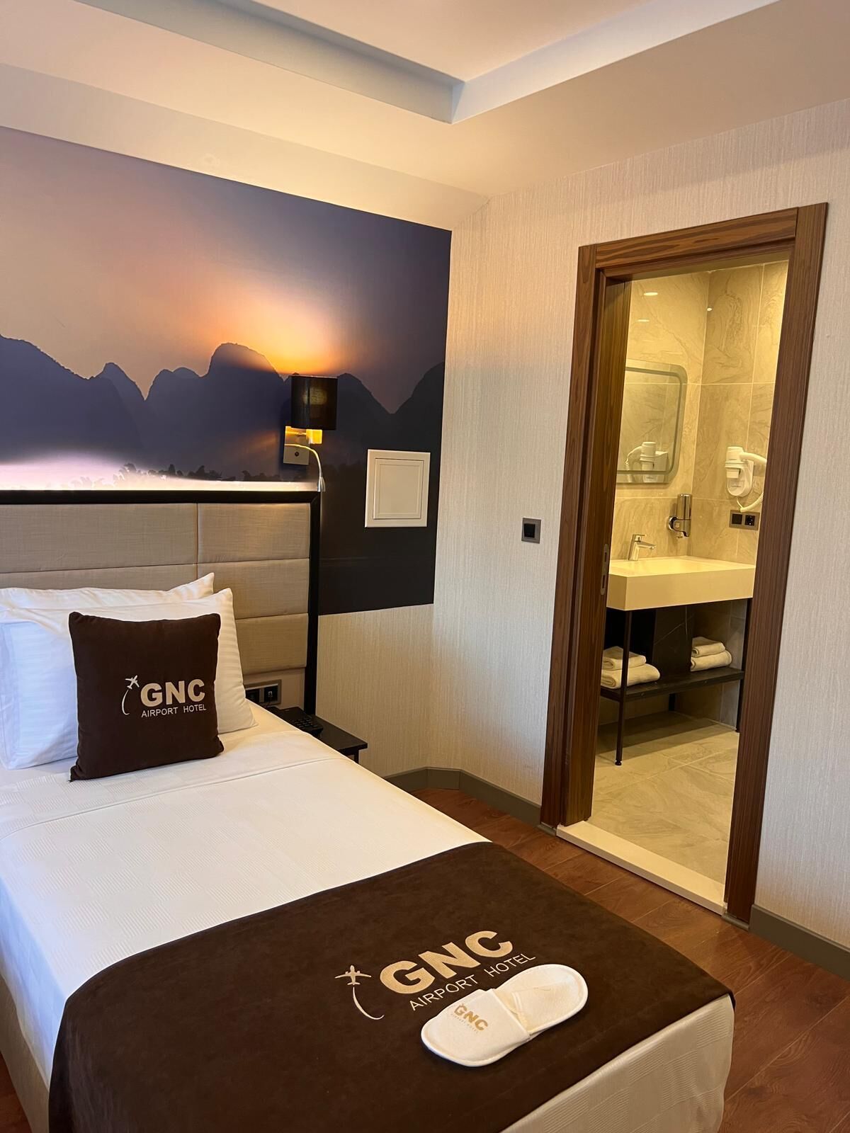 A hotel room with a bed, a nightstand, a wall-mounted lamp, a bathroom with a mirror and towel rack, and a GNC logo on the bed and nightstand.