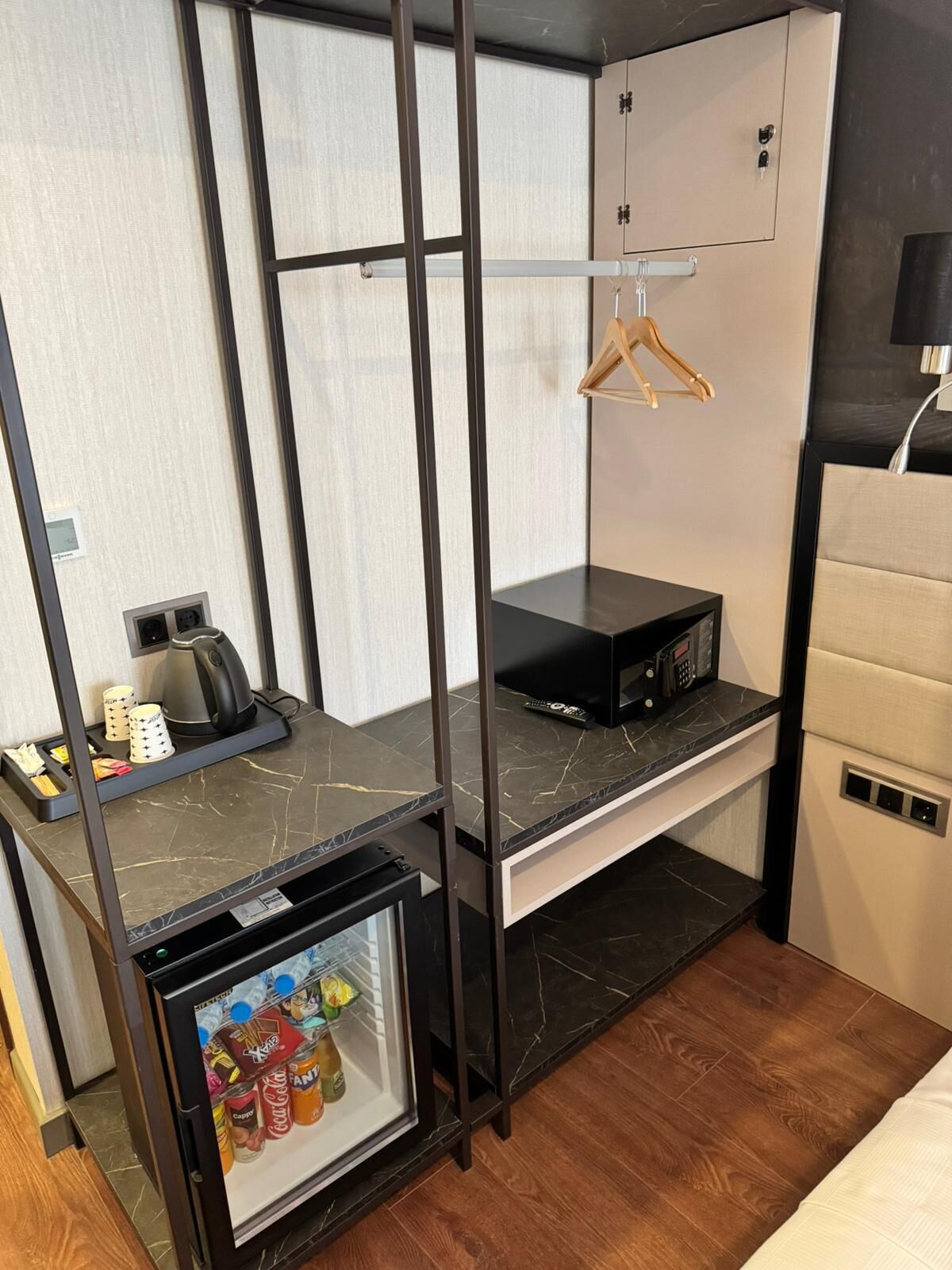 A compact hotel room with a kitchenette, a minibar, a small refrigerator, a wooden hanger, and a bed.