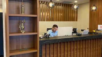 A reception desk with a wooden panel design, a man seated behind it, and a shelf with various decorative items.