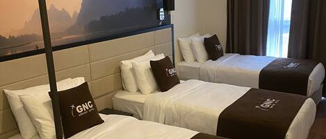 A hotel room with three beds, each with a GNC Airport Hotel pillow, a headboard, and a wall-mounted lamp.