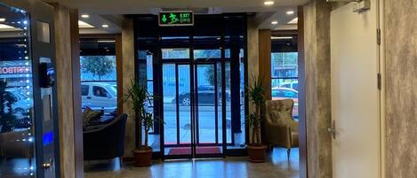 A modern hotel lobby with a wooden ceiling, a reception desk, and a glass door leading to the parking area.
