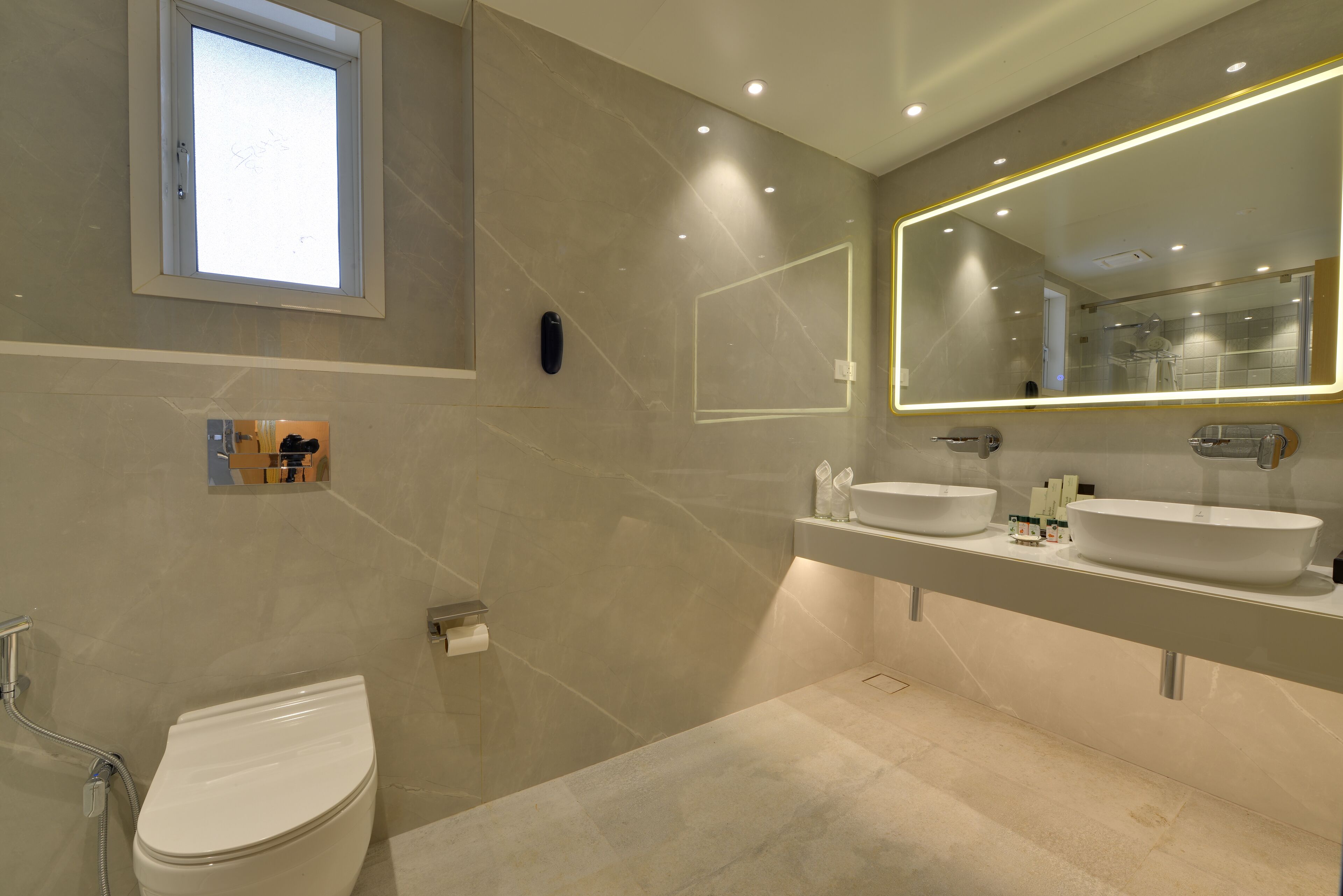A modern bathroom with a toilet, two sinks, a large mirror, and a window.
