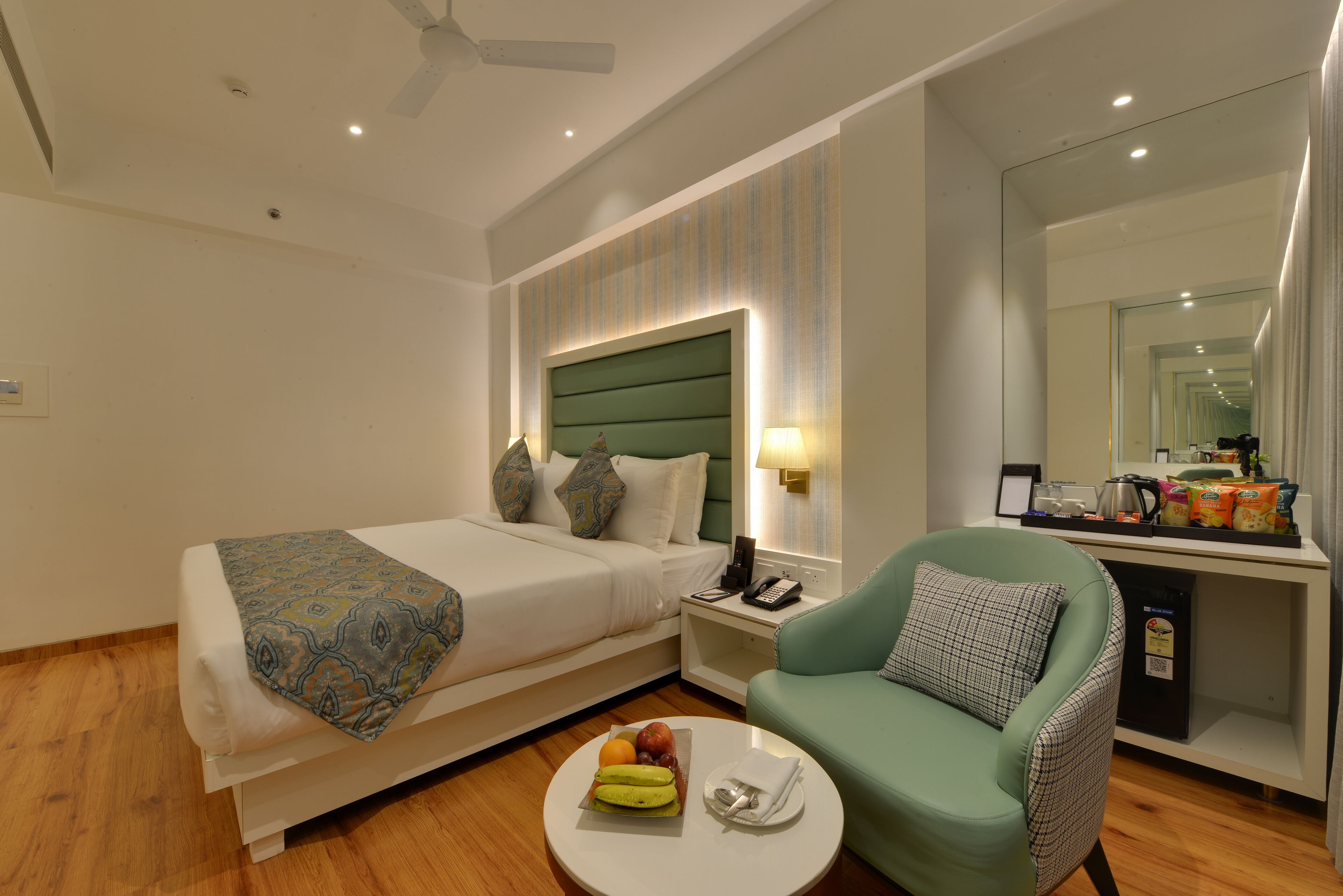 Una habitación de hotel con una cama, un sillón verde, una mesita con fruta, una mesita de noche con un teléfono y un espejo.