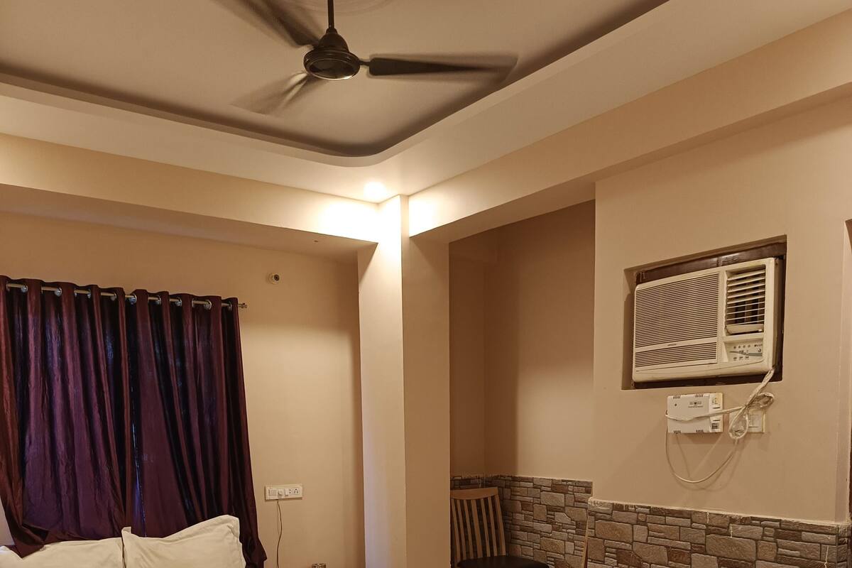 A bedroom with a bed, a ceiling fan, a window with an air conditioner, a chair, and a small table.