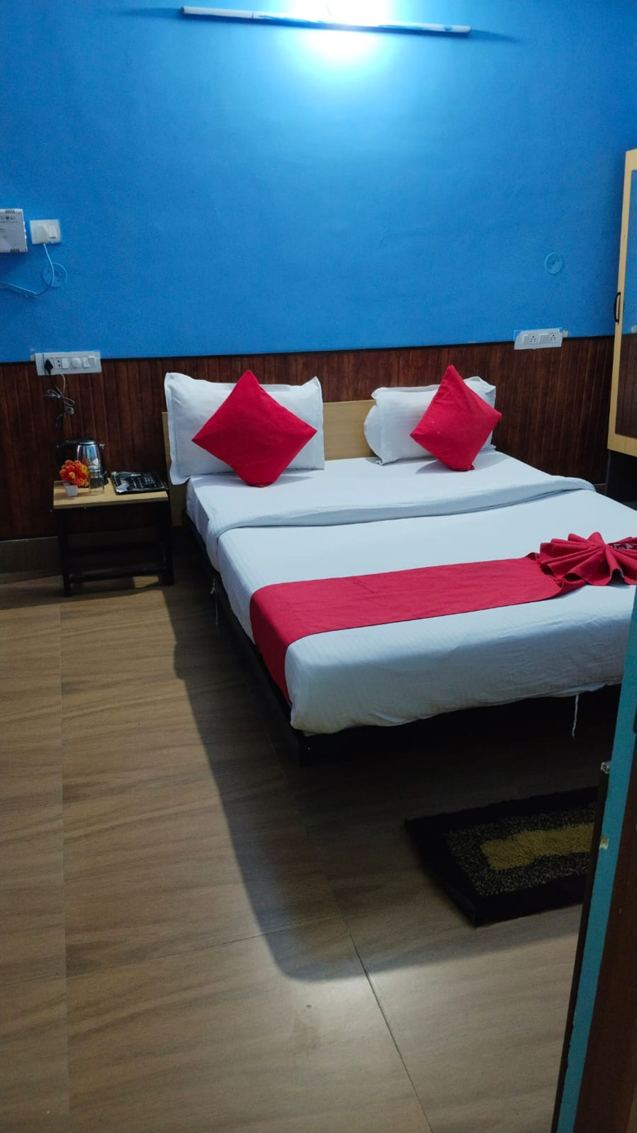Hotelkamer met een groot bed, twee rode kussens, een witte dekbedovertrek en een rode deken. Er staat een nachtkastje met een lamp en een klein tafeltje met een bloevas. De kamer heeft blauwe wanden en houten lambrisering in het onderste deel.