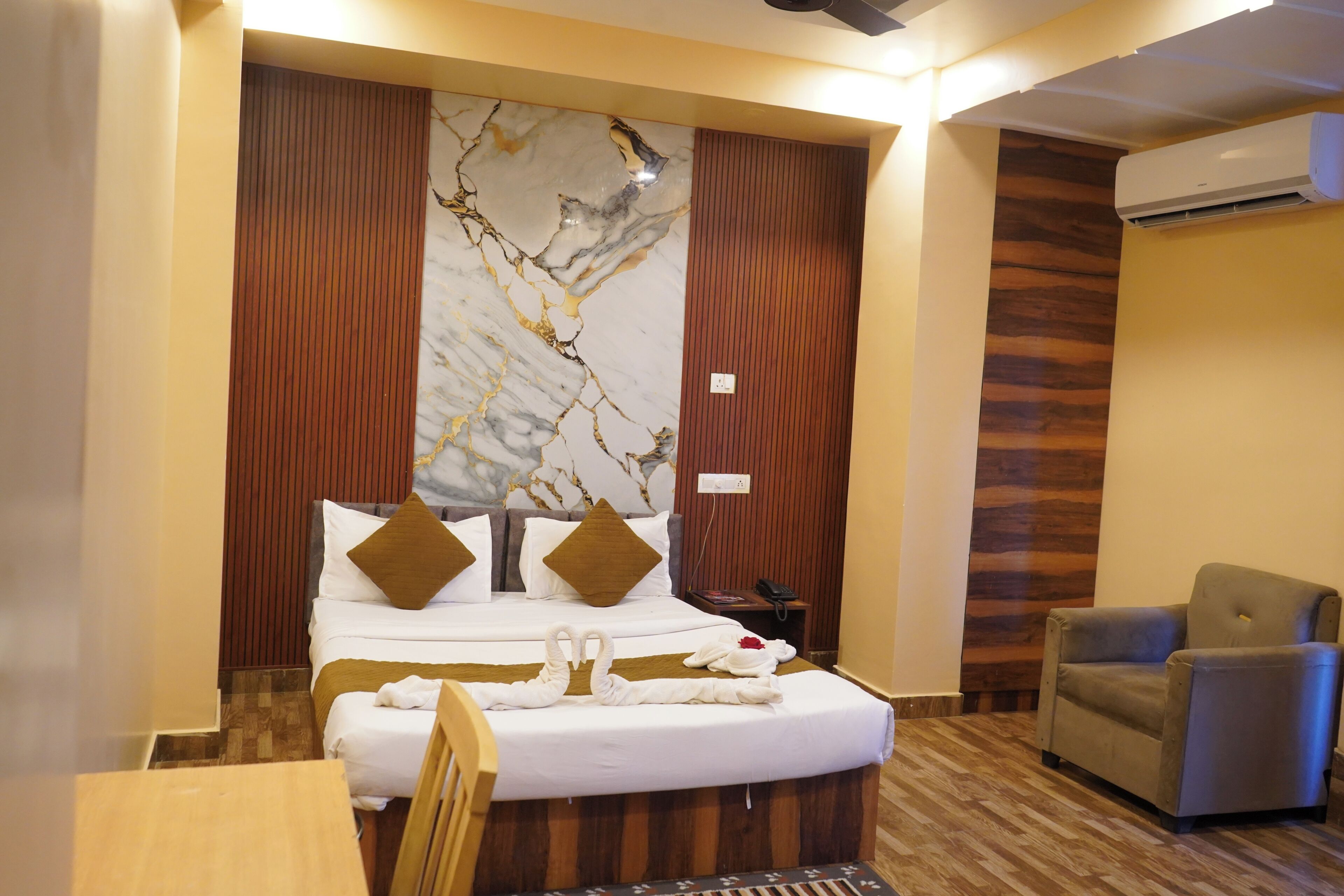 Une chambre d’hôtel avec un grand lit, une tête de lit en bois, un mur en marbre, un plancher en bois, un fauteuil gris et une petite table.